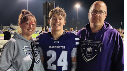 Three people smiling and posing together at a nighttime outdoor sports event, with one person wearing a football uniform and the others wearing hoodies. Description generated by AI