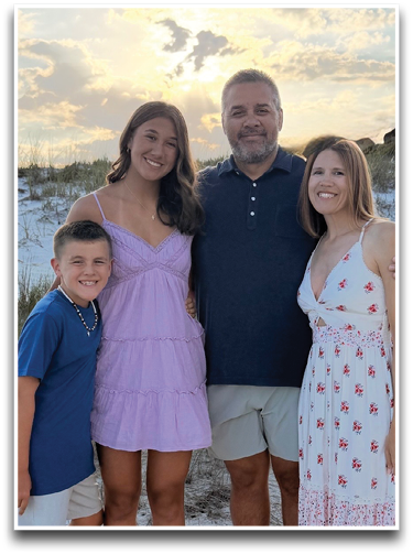 Four people standing close together on a sandy beach at sunset with one person wearing a blue shirt and shorts, one in a light purple dress, one in a dark polo shirt and light shorts, and one in a white dress with red floral patterns. Description generated by AI