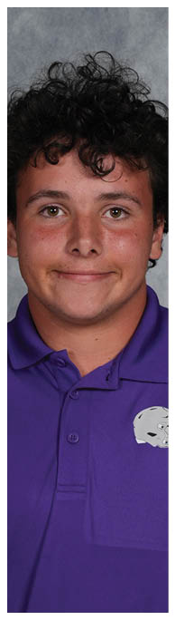 A person with curly hair wearing a purple collared shirt with a small white logo is smiling against a gray mottled background. Description generated by AI