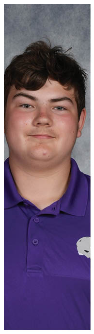 A person with short curly hair wearing a purple collared shirt is standing against a gray mottled background. Description generated by AI