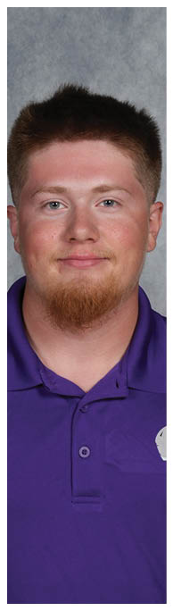 A person with short hair and a goatee is wearing a purple collared shirt and smiling against a gray background. Description generated by AI