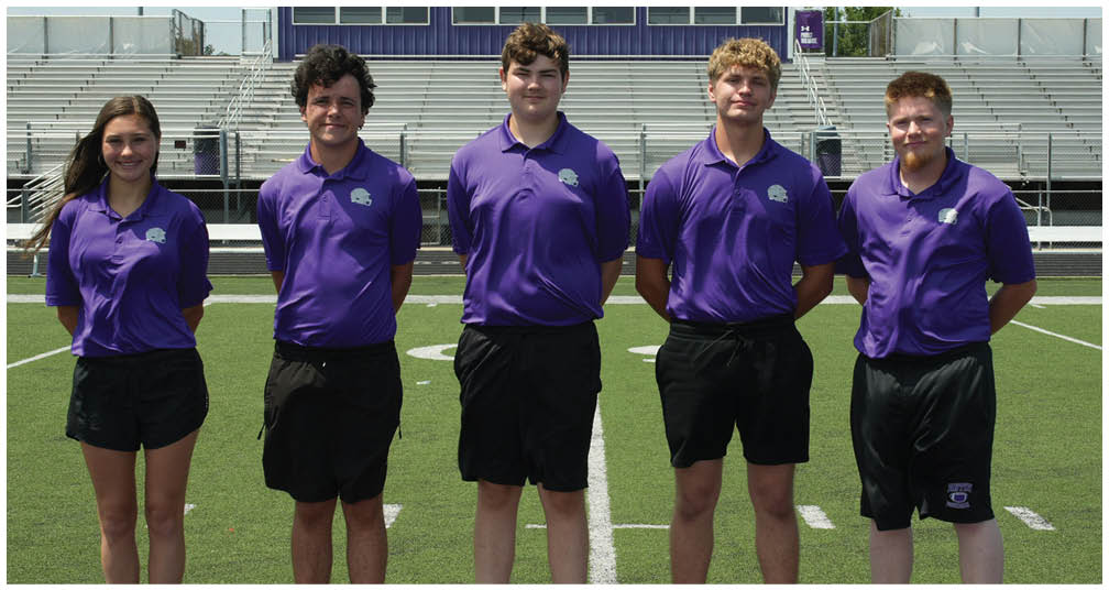 Five people wearing matching purple polo shirts and black shorts stand side by side on a sports field with empty bleachers in the background. Description generated by AI
