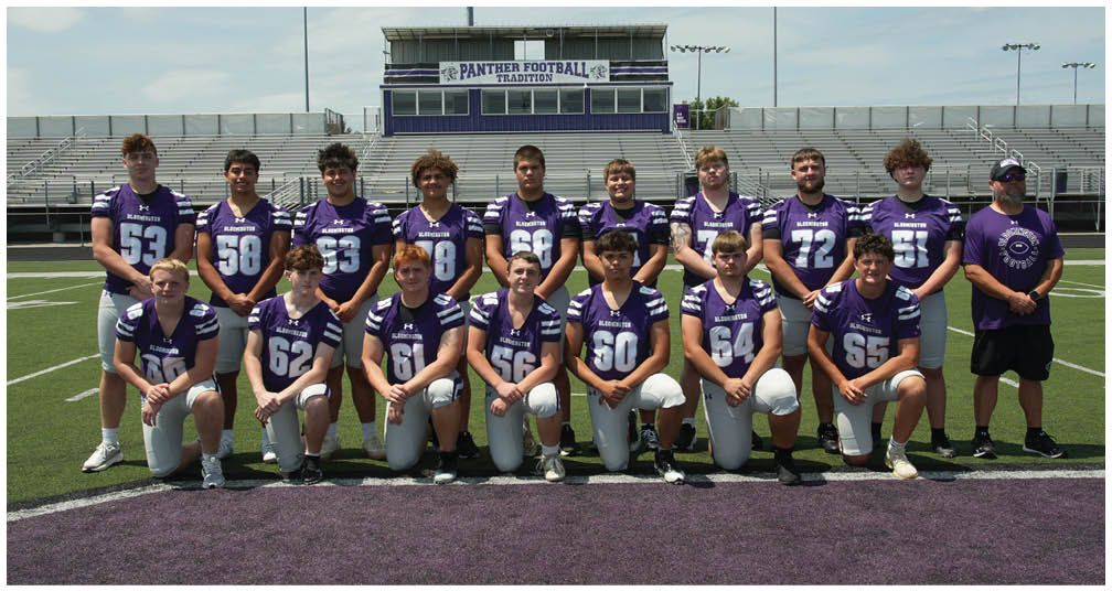 A group of people wearing purple and white football uniforms pose together on a football field with bleachers and a scoreboard in the background. Description generated by AI