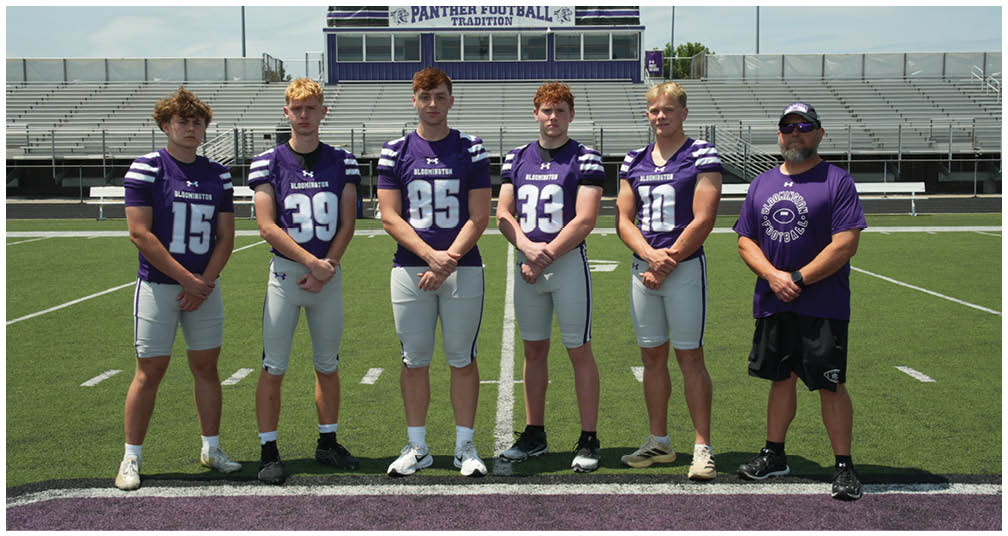 A group of people wearing purple and white football uniforms pose together on a football field with bleachers and a scoreboard in the background. Description generated by AI