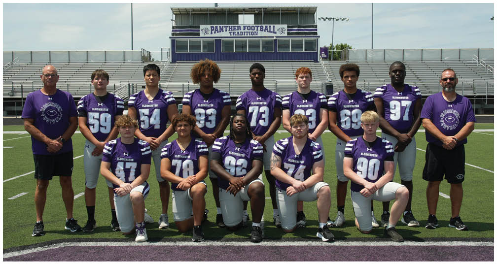 A group of people wearing purple and white football uniforms pose together on a football field with bleachers and a scoreboard in the background. Description generated by AI