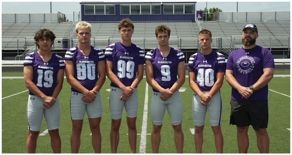 A group of people wearing purple and white football uniforms pose together on a football field with bleachers and a scoreboard in the background. Description generated by AI
