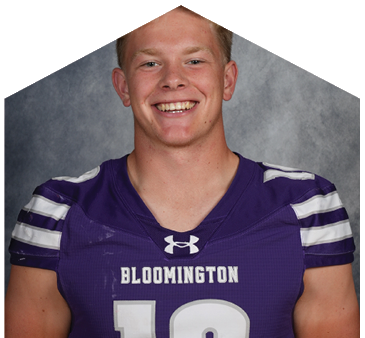 A person with short light hair is smiling and wearing a purple sports jersey with white stripes on the shoulders against a gray mottled background. Description generated by AI
