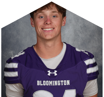 A person with short tousled hair is smiling and wearing a purple sports jersey with white stripes on the shoulders against a gray mottled background. Description generated by AI