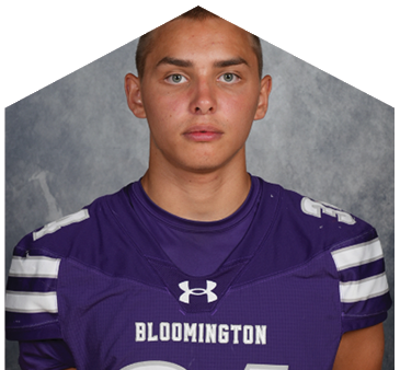 Person wearing a purple Bloomington sports jersey with white stripes on the sleeves posing against a gray backdrop. Description generated by AI