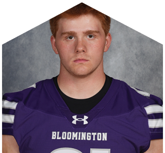 A person with short hair wearing a purple sports jersey with the word Bloomington on the front is looking directly at the camera against a gray background. Description generated by AI