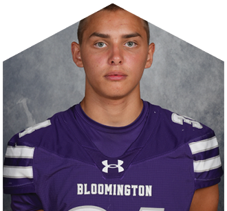 Person wearing a purple Bloomington sports jersey with white stripes on the sleeves posing against a gray backdrop. Description generated by AI