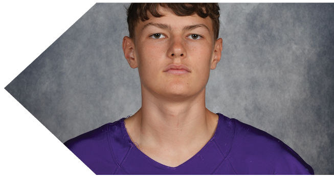 A person with short curly hair wearing a purple sports jersey is looking directly at the camera against a gray textured background. Description generated by AI