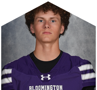 A person with curly hair wearing a purple Bloomington sports jersey with white stripes on the sleeves is looking straight ahead against a gray background. Description generated by AI