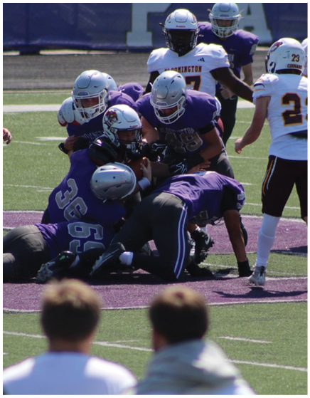 Several people in football uniforms are engaged in a tackle on a field while two others stand nearby watching. Description generated by AI