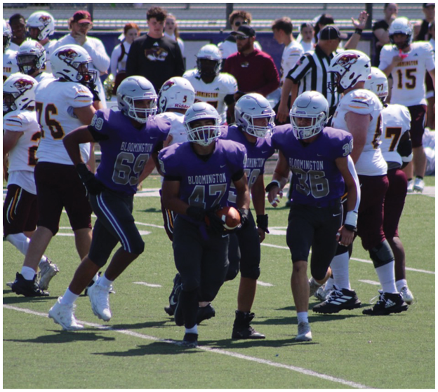 Adults wearing football uniforms are playing on a field with one player in a purple jersey running with the ball while others in white and purple jerseys are positioned around. Description generated by AI