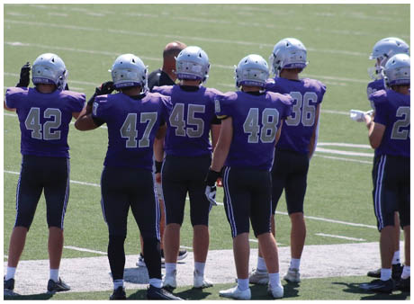 Several people wearing dark sports uniforms with numbers stand on a grassy field near a white sideline. Description generated by AI