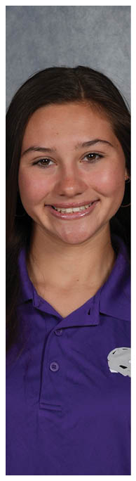 A person wearing a purple shirt stands against a dark textured background with their arm bent behind their back. Description generated by AI