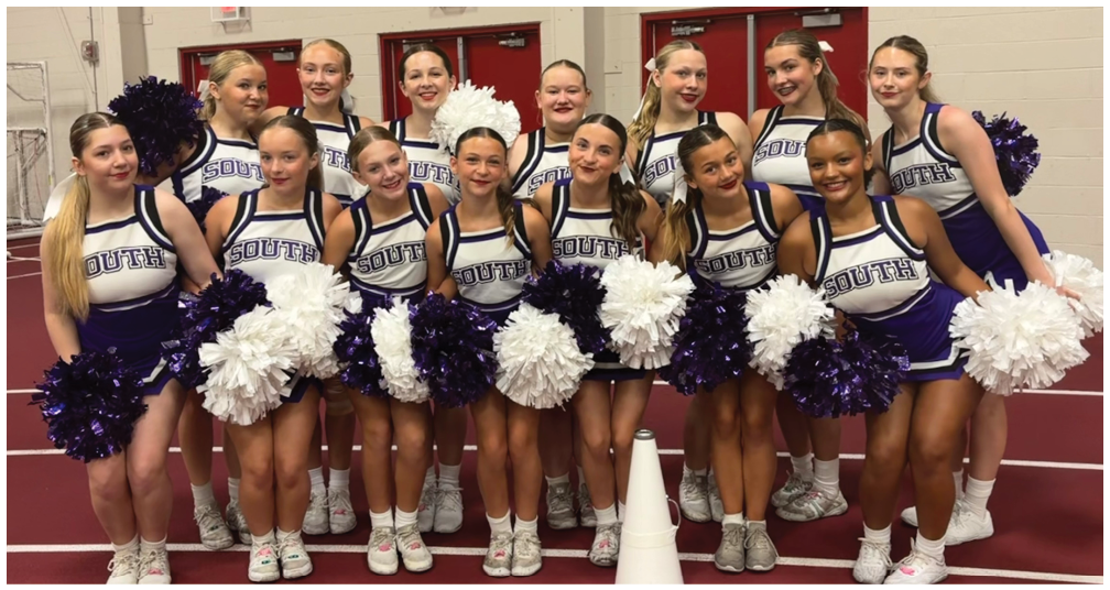 A group of people dressed in matching cheerleading uniforms holding white and purple pom-poms are standing closely together indoors. Description generated by AI