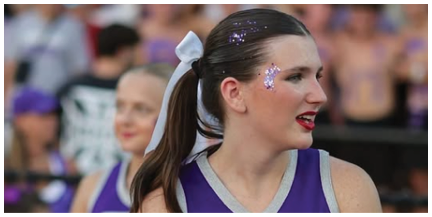 A person with a ponytail tied with a white ribbon and glitter on their face is in focus with a blurred crowd in the background. Description generated by AI