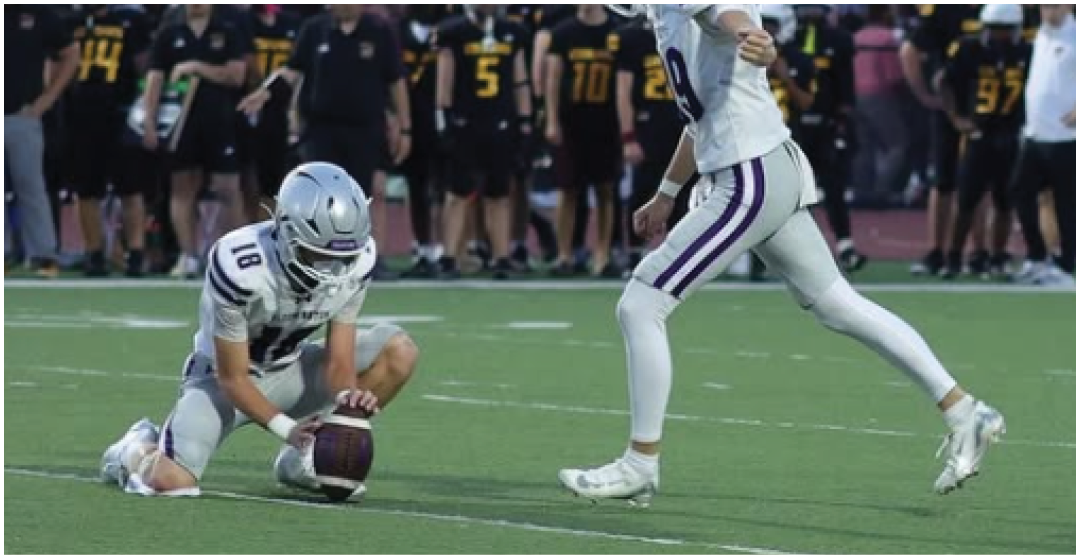 Two people in football uniforms are on a field with one holding the ball on the ground and the other preparing to kick it. Description generated by AI
