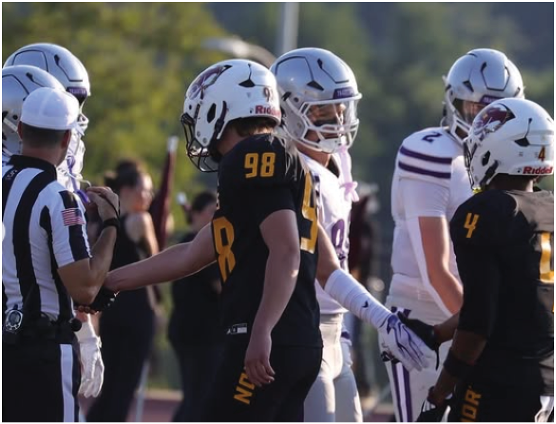 Several people wearing football helmets and uniforms are gathered outdoors, with some wearing black and yellow and others in white and purple jerseys. Description generated by AI