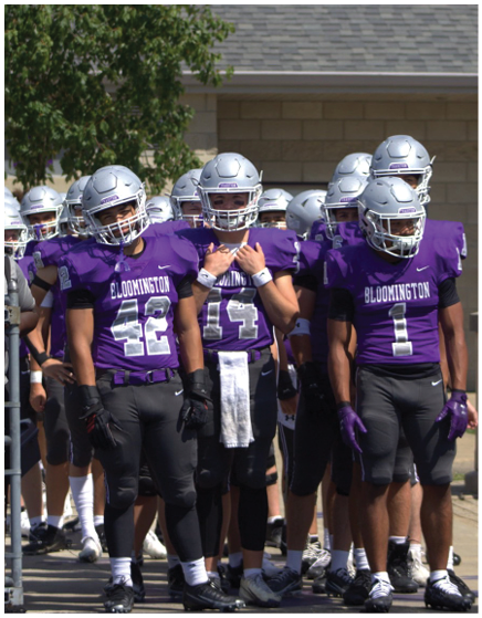 A group of people wearing purple football uniforms and silver helmets stand together outdoors on a sunny day. Description generated by AI