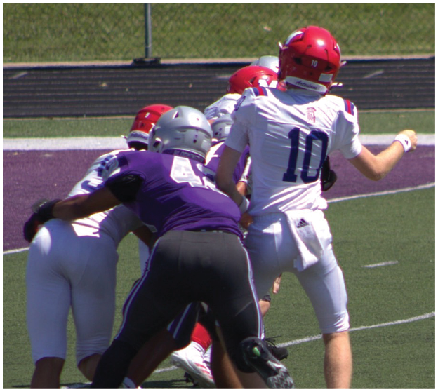 A group of people wearing football uniforms are engaged in a play on a football field with one person in a white jersey preparing to throw a ball. Description generated by AI