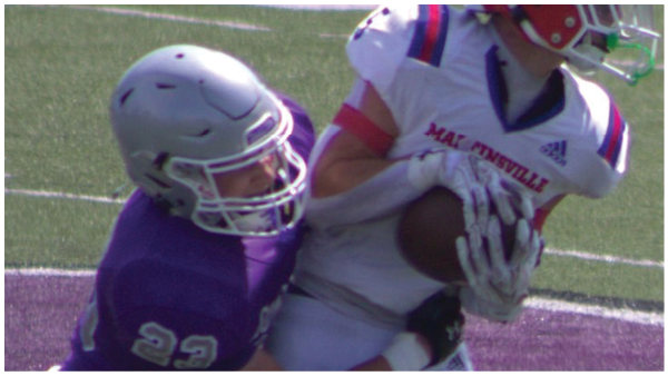 Two people wearing football helmets and uniforms are engaged in a play on a grassy field, with one person holding the ball while the other attempts to tackle. Description generated by AI