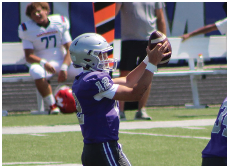 A person wearing a purple football uniform and helmet is catching a football on a field with other players and a sideline in the background. Description generated by AI