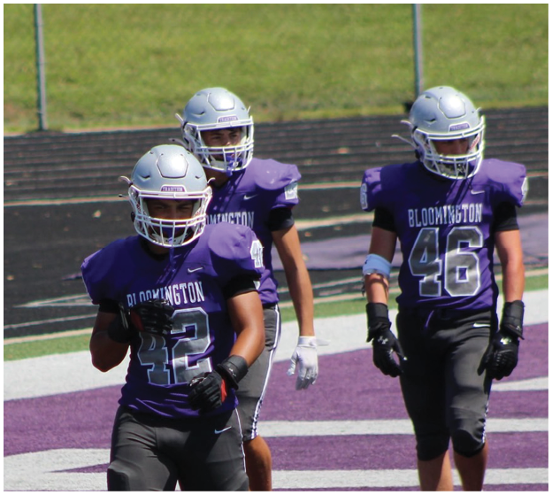 Three people wearing purple Bloomington football uniforms and silver helmets are standing on a field with a purple and white end zone. Description generated by AI