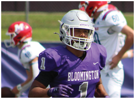 A person wearing a purple Bloomington football uniform and white helmet is on a field with two other players in white uniforms and red helmets in the background. Description generated by AI