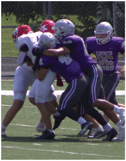 Four people in football uniforms are engaged in a tackle on a grassy field with a chain-link fence and trees in the background. Description generated by AI