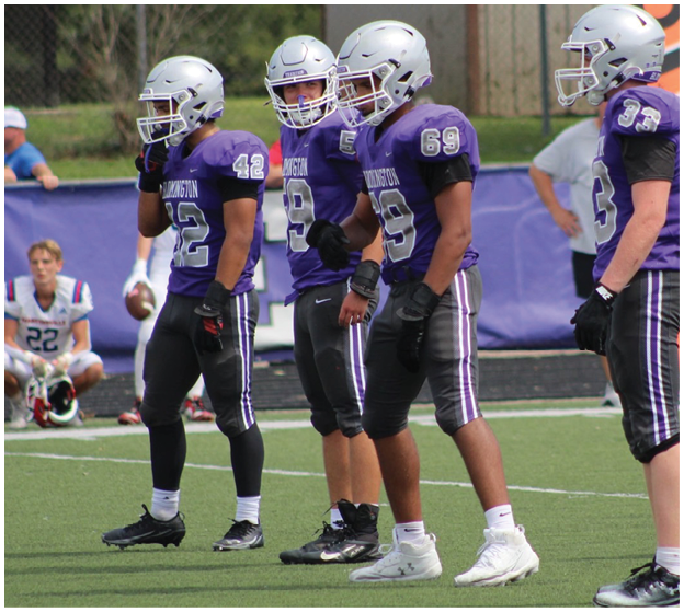Four people wearing purple football uniforms and helmets stand on a football field during a game or practice. Description generated by AI