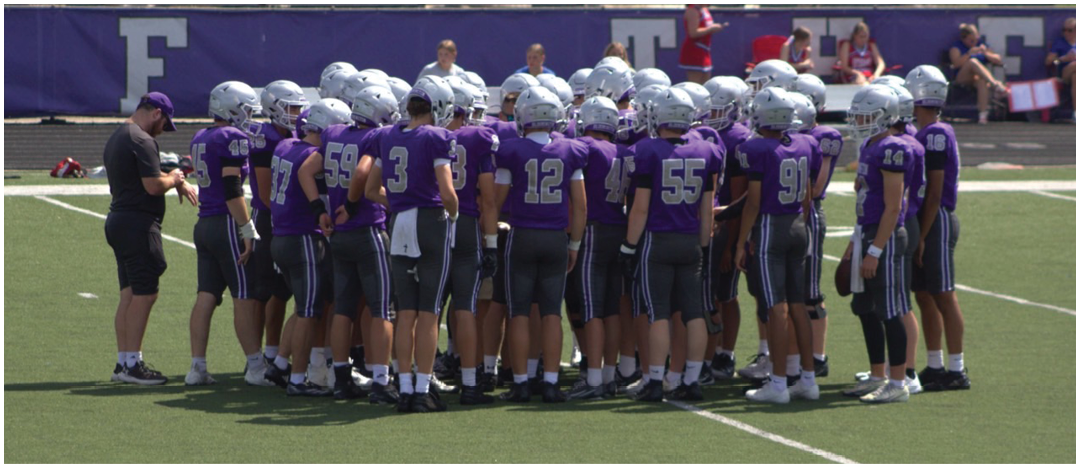 A group of people wearing purple sports jerseys and silver helmets are huddled together on a football field. Description generated by AI