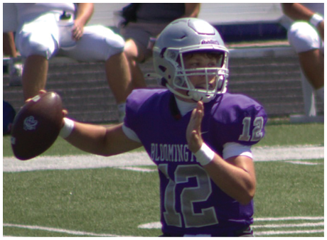 A person wearing a purple football uniform with the number 12 is preparing to throw a football on a field with other players and spectators in the background. Description generated by AI