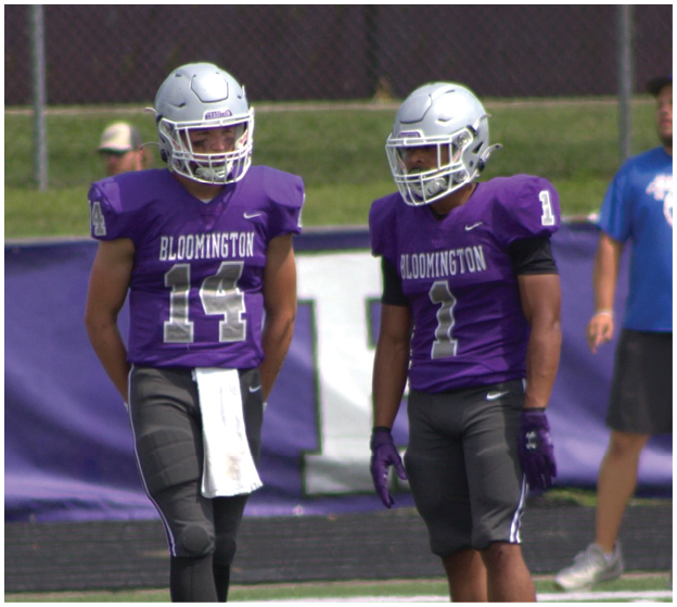 Two adults wearing purple Bloomington football uniforms and helmets stand on a field with a blurred person in the background. Description generated by AI