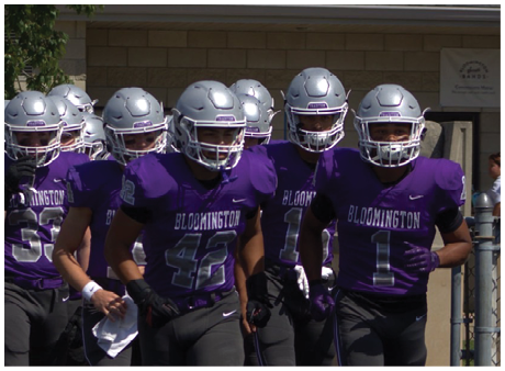 Four people wearing purple Bloomington football uniforms and silver helmets stand closely together outdoors. Description generated by AI
