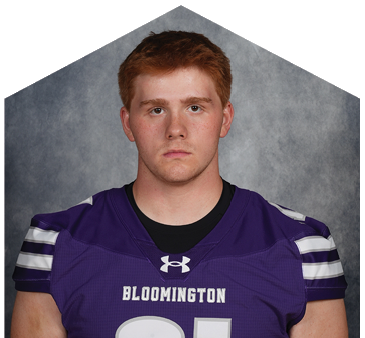 Person with short hair wearing a purple Bloomington sports jersey looking directly at the camera against a gray background. Description generated by AI