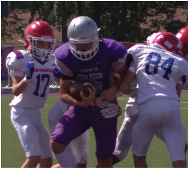 Four people in football uniforms are engaged in a play on a grassy field with one person in a purple uniform holding the ball while three others in white uniforms with red helmets attempt to block or tackle. Description generated by AI