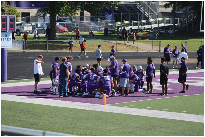 A group of people in purple sports uniforms and casual clothing are gathered on a field near a scoreboard with others walking and standing in the background near a fence and parked cars. Description generated by AI