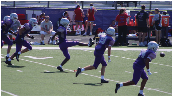 Four people in purple football uniforms and helmets are on a field with one kicking a football while others run forward, and several people are in the background near the sideline. Description generated by AI