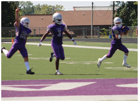 Three people in purple football uniforms and helmets are on a football field with one person running while holding a football and the other two celebrating. Description generated by AI