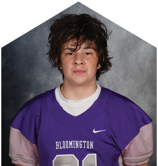 A person with short curly hair wearing a purple sports jersey with white stripes and the word Bloomington on the front is looking directly at the camera against a gray background. Description generated by AI
