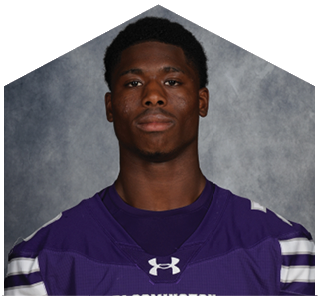 A person wearing a purple Bloomington sports jersey with white stripes on the sleeves is standing against a gray textured background. Description generated by AI
