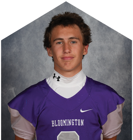 A person with short curly hair wearing a purple sports jersey with white stripes and the word Bloomington on the front is looking directly at the camera against a gray background. Description generated by AI