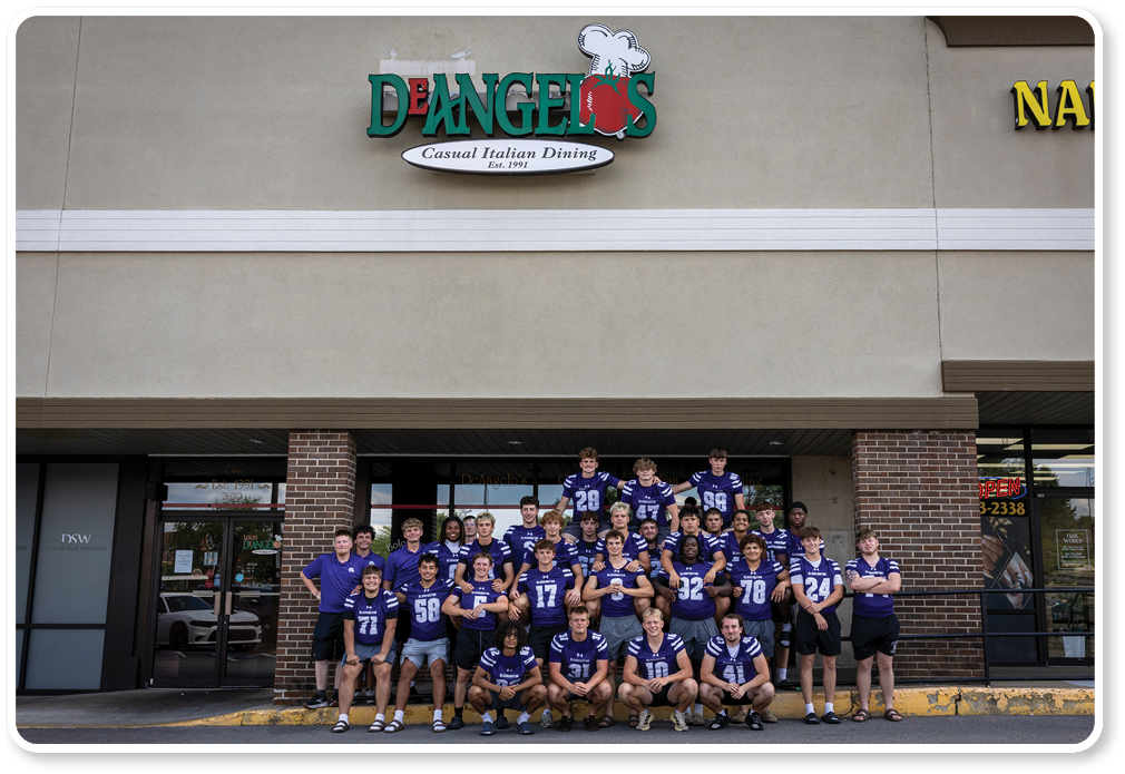 A group of people wearing matching blue sports jerseys pose together outside a restaurant named D'Angelo's. Description generated by AI