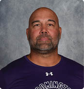 A person with a shaved head and beard wearing a purple Under Armour shirt with the word Bloomington and a football graphic on it stands against a gray mottled background. Description generated by AI