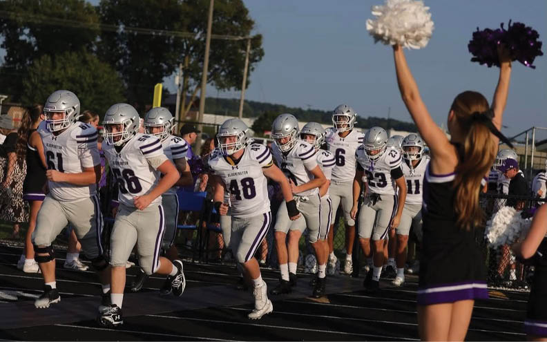 Four people wearing football helmets and white jerseys with purple stripes are standing outdoors on a field. Description generated by AI