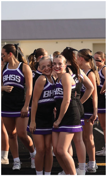 A group of people in matching purple and black cheerleading uniforms are standing together outdoors, with two smiling individuals posing closely in the foreground. Description generated by AI