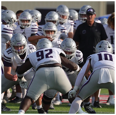 A group of football players in helmets and uniforms are lined up on a field preparing for a play with a coach standing nearby. Description generated by AI